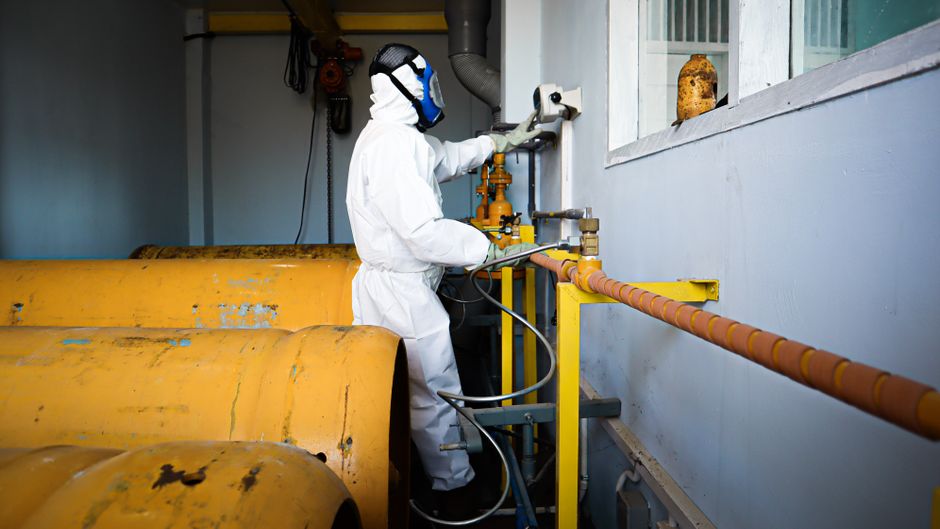Worker in full protective suit and gas mask handles industrial pipes indoors in Indonesia.